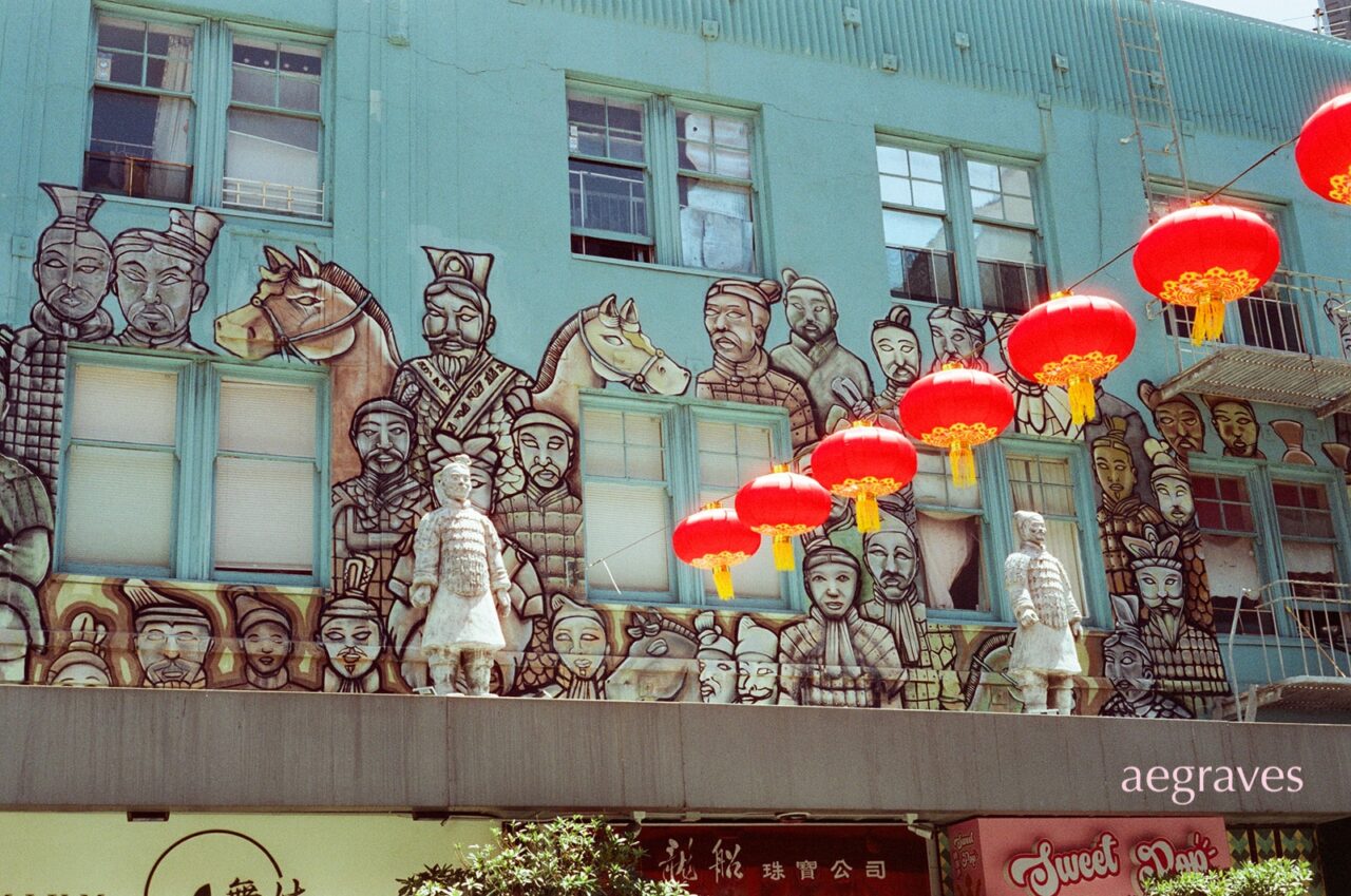 Glowing red lanterns hang in front of soldier sculptures and a related mural with a pale green background in SF Chinatown