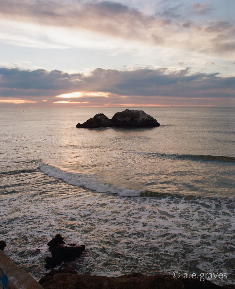 A view of rocks offshore in the Pacific Ocean during a cloudy sunset; rocks in the foreground suggest the view is taken from land