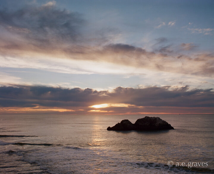 View of the evening horizon with clouds, the Pacific Ocean, a prominent two peak rock, and shallow foreground waves
