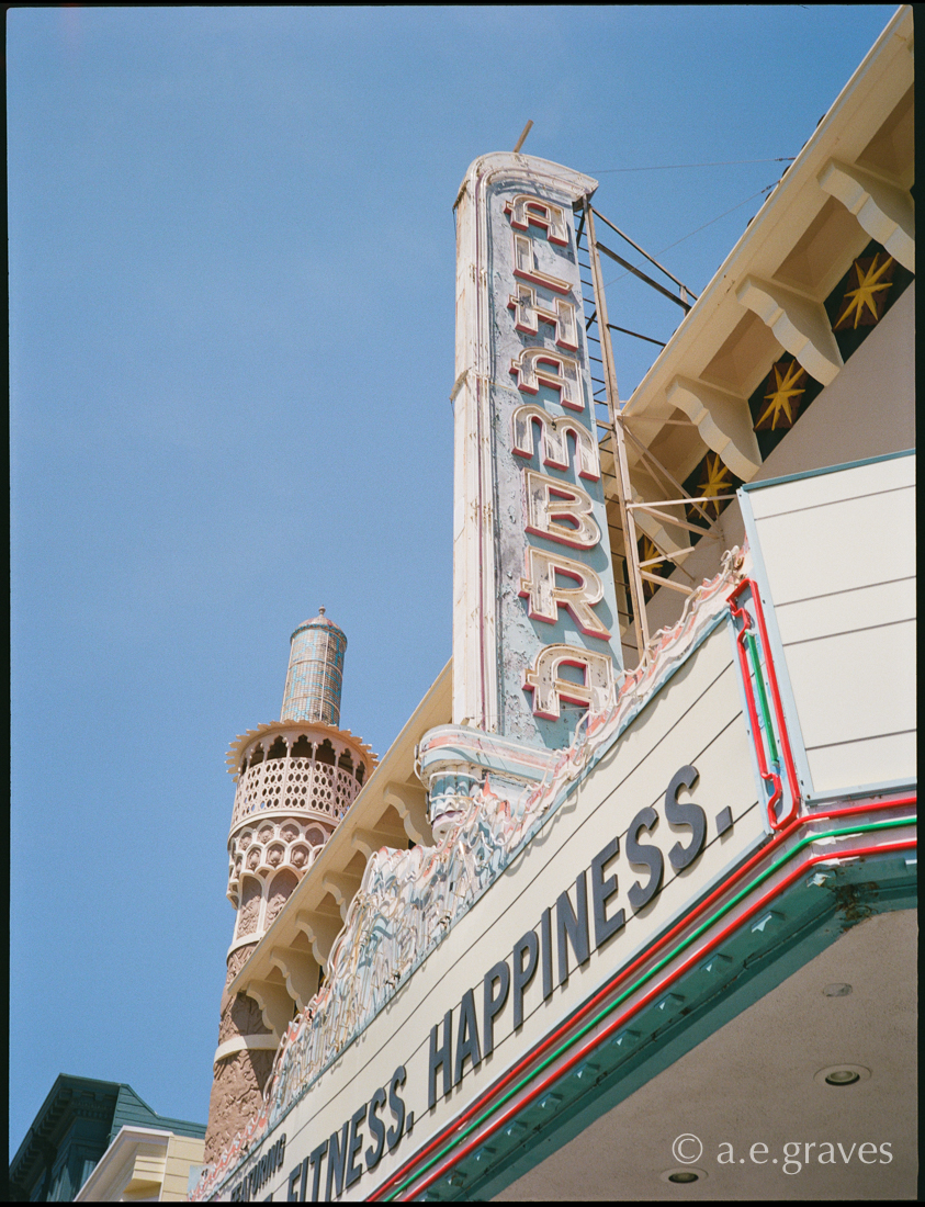 Front upward view of the pastel blue and pink Alhambra neon marquee, with a partial minaret in the background, and the word HAPPINESS on the sign board