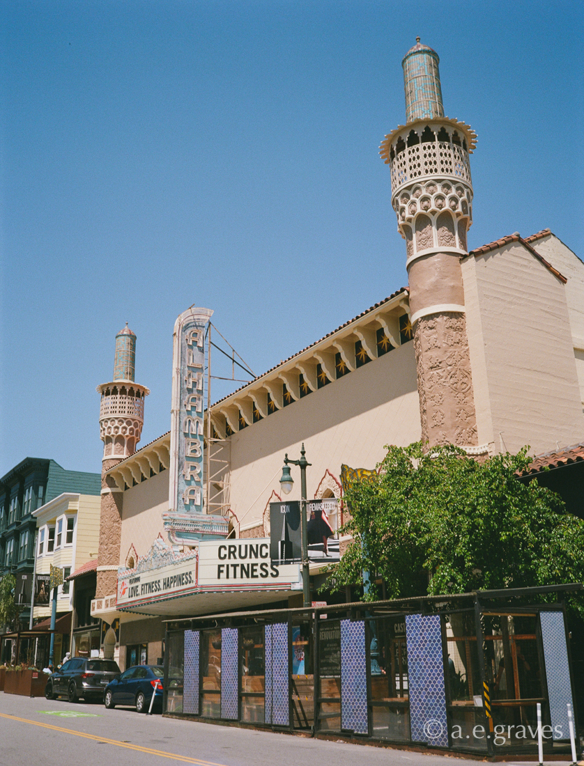 Street view of the entire, wide front of the Alhambra Theater from the distance down the street, featuring both minarets, the neon sign, and the signboard. A structure in the street obscures the storefront