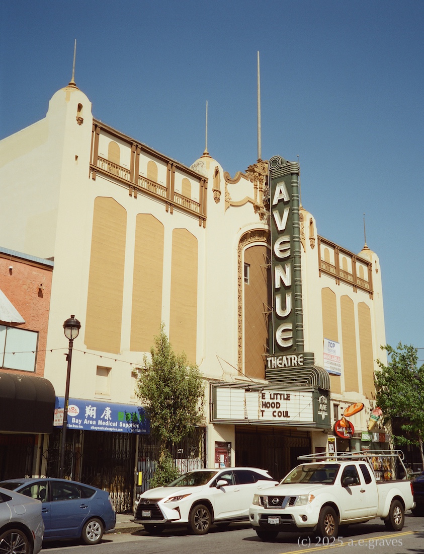Street view of a cream and ochre movie theater with darker bronwn decorations, and a tall, green theater marquee with neon letters reading AVENUE