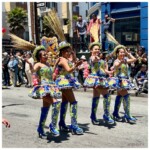 Four dancers in elaborately beaded blue, green, yellow, and orange costumes, with matching high-heeled boots and small hats with taller straw decorations.