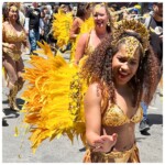 Dancers in gold bikinis, crowns, and yellow feathers smiling both generally and at cute children in the crowd