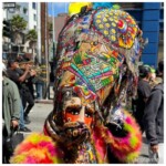 Masked character with an elaborately sewn and beaded headdress of many colors.