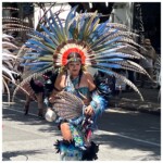 Dancer with blue-themed feathered costume referencing Central American cultures; predominantly featuring blue feathers, embroidery, and jewelry, with brown, cream, and red-orange details and a striped feather fan.