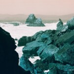 View of turquoise rocks under a pale orange sky with the Pacific Ocean and distant hills in the background