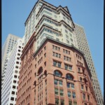 View looking up at a highrise with a red brick building topped with a narrower yellow highrise with dark protruding shades