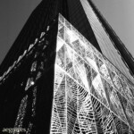 This film displays strong contrast, white-hot reflections, and deep shadows Monochrome image of a skyscraper corner; a screen looking like a segmented topographical map dominates the image, its right side reflecting hotly.
