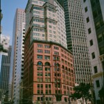 Color image of the deYoung building at Market and Kearney, with the brick red structure in the lower half and a butter yellow addition above, surrounded by other, less colorful highrises