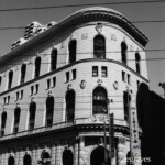 The film has pleasant midtones, and the shadows preserve detail. There is less detail in the highlights than Ilford film. Monochrome image of a rounded, historic-looking building with overhanging cornices and rows of arched windows with one row of smaller, rectilinear windows. Wires cross the image horizontally.