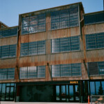 Sunlit color image of a four story building with a glass ground floor and metal and glass upper floors display rust and large windows