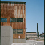 View of an industrial building corner, with rusted panels of different densities of deep red; a gray boxy building sits in the lower left; a street turns the corner to the right of the building; other warehouses and a smokestack are visible in the distance