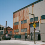 View of a tall building with variegated rust patterns on the upper reaches of the tall wall; a metal fence and streetlight with a left turn sign are in the foreground; other buildings and a smokestack are visible in the background