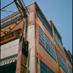 Color image looking up at a rusted industrial building, with an incomplete scaffold-like steel canopy and its support members in the foreground