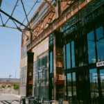 A tall series of window reflect nearly teal in front of steel beams holding up an unseen roof structure; rusty sheet metal panels cover the upper portions of the building