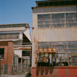 Two story industrial building with four yellow utility bells suspended from the wall, casting shadows; another warehouse with utility piping connecting to the main building is visible to the left