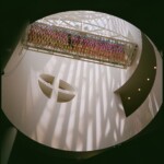 Color image looking up into SFMoMA's Botta atrium, with a person crossing the bridge and sunlight casting long shadows down the walls beneath Eliasson's colorful tunnel sculpture