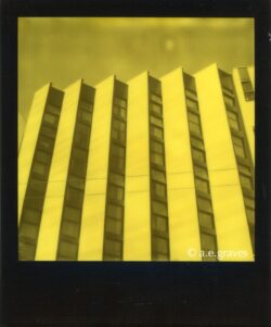 Monochrome black and yellow Polaroid image of a tall building facade with a fan-fold structure of blank walls facing left and windows facing right